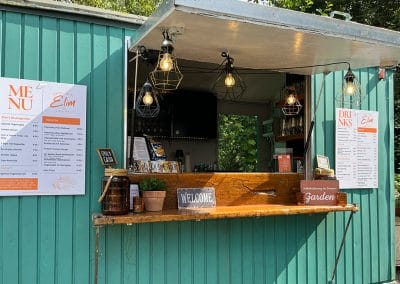 Die Verkaufstheke des Sommergartens „Kreativwerk Elim“ in einem grünen Bauwagen mit Menü- und Getränkekarten an der Außenwand.