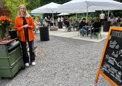 Eine lächelnde Mitarbeiterin in einem orangefarbenen Hemd im Außenbereich des Sommergartens mit Gästen, Sonnenschirmen und einer Kreidetafel im Vordergrund.