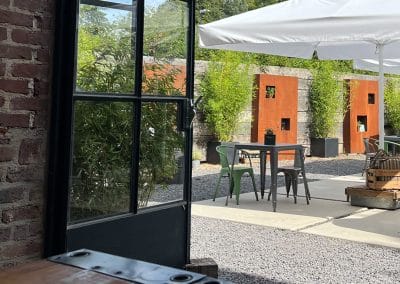Blick durch ein geöffnetes Sprossenfenster auf den Kiesplatz des Sommergartens mit modernen Metalltischen und großen weißen Sonnenschirmen.