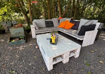 Gemütliche Lounge-Ecke aus weiß gestrichenen Paletten mit grauen Polstern und orangefarbenen Kissen unter schattigen Bäumen.
