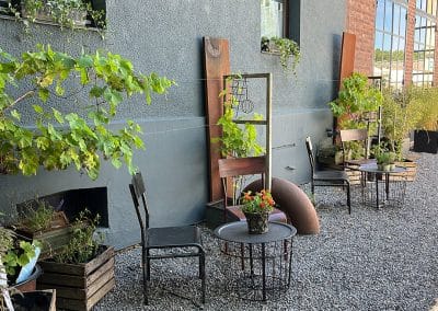 Schlichte, stilvolle Sitzgelegenheiten mit schwarzen Metalltischen und Stühlen auf grauem Kies vor einer modernen Fassade mit Kletterpflanzen.