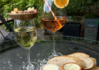 Zwei Weingläser – eines mit Weißwein, eines mit einem orangefarbenen Spritz-Drink – auf einem Glastisch mit Brot und Kräuterbutter.