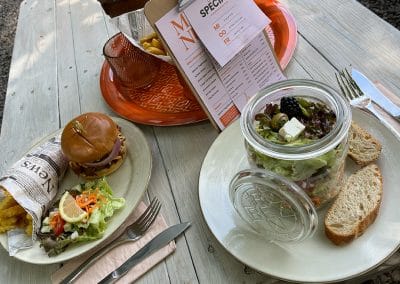 Ein gedeckter Tisch mit einem Burger in Zeitungspapier, Pommes und einem Schichtsalat im Glas neben einer Menükarte auf einem Klemmbrett.